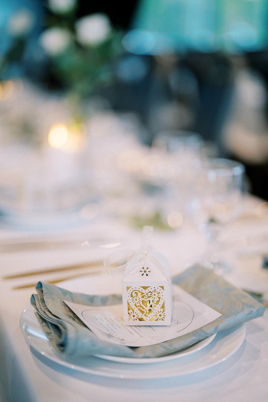 Elegant wedding table setting with a decorative white and gold favor box on a folded napkin and plate, surrounded by softly blurred glassware and flower decor in the background.