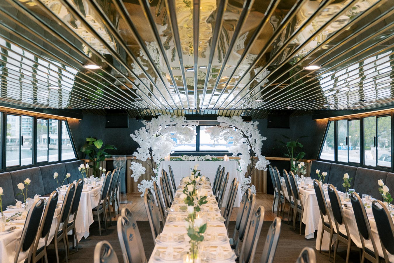 Elegant dining setup on a luxury river cruise boat featuring long tables with white tablecloths, neatly arranged tableware, and floral centerpieces, complemented by large windows offering scenic views.