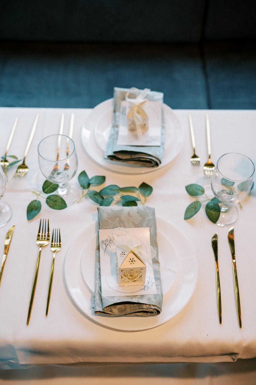 Elegant table setting with gold cutlery, white plates, blue napkins, and small decorative gift boxes on a white tablecloth, enhanced by green leaf accents and clear water glasses for a stylish dining ambiance.