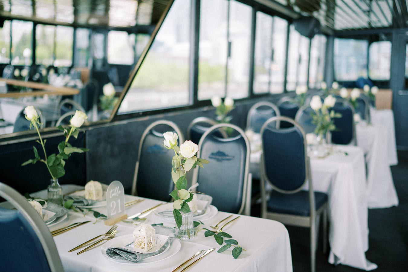 Elegant dining setup on a boat with white tablecloths, blue chairs, and delicate white rose centerpieces, creating a serene and sophisticated ambiance for a special event or celebration.