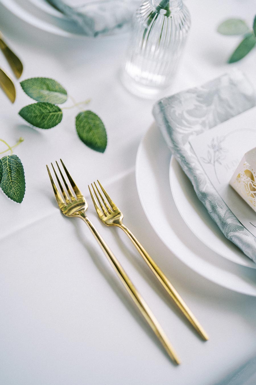 Elegant table setting featuring a gold fork and salad fork on a white tablecloth, accompanied by green leaves, a textured blue napkin, a delicate lace gift box, and a clear glass vase for a sophisticated dining experience.