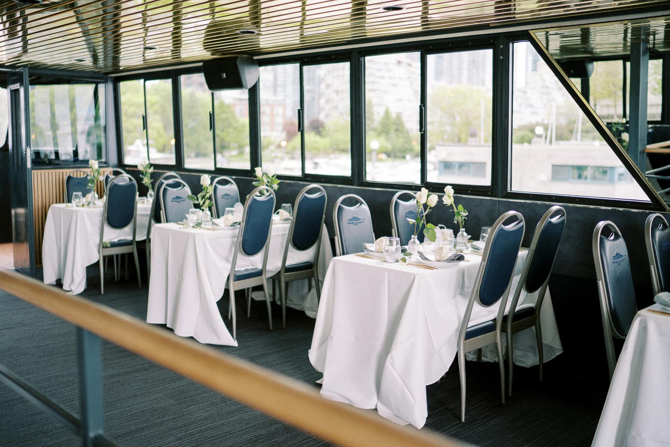 Elegant dining area on a boat with white tablecloths and blue chairs, set for a formal event with windows showcasing a view of greenery outside.