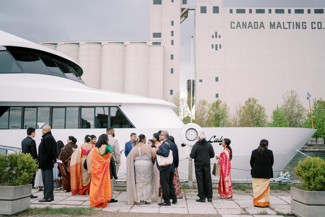 Group of people in colorful traditional attire gathered on a pier beside a large white yacht, with the Canada Malting Co. building and greenery in the background.