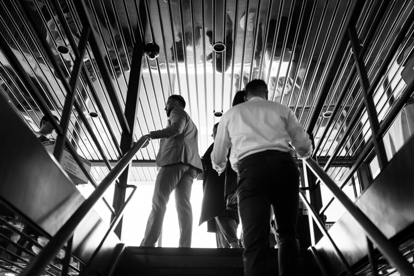 Black and white image of people in business attire walking up a modern staircase, with a reflective ceiling above, conveying an urban corporate environment.