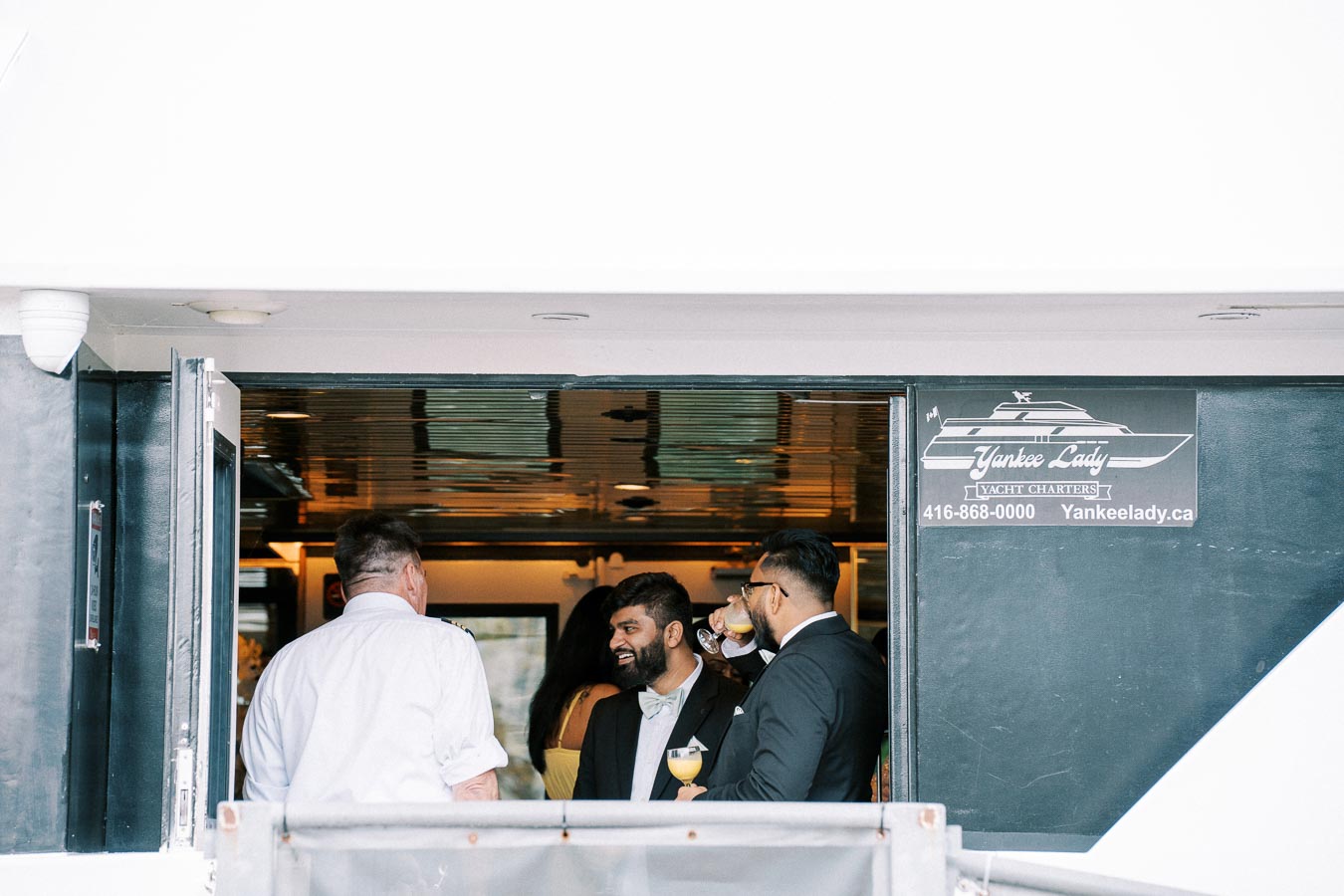Group of people socializing inside a yacht, with formal attire and drinks in hand, under the sign for Yankee Lady Yacht Charters.
