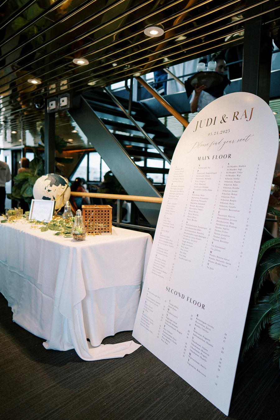 Wedding reception seating chart for Judi and Raj on May 24, 2025, displayed on a large white board. The setup includes a decorated table with a globe and ambient lighting in a modern venue with stairs in the background.