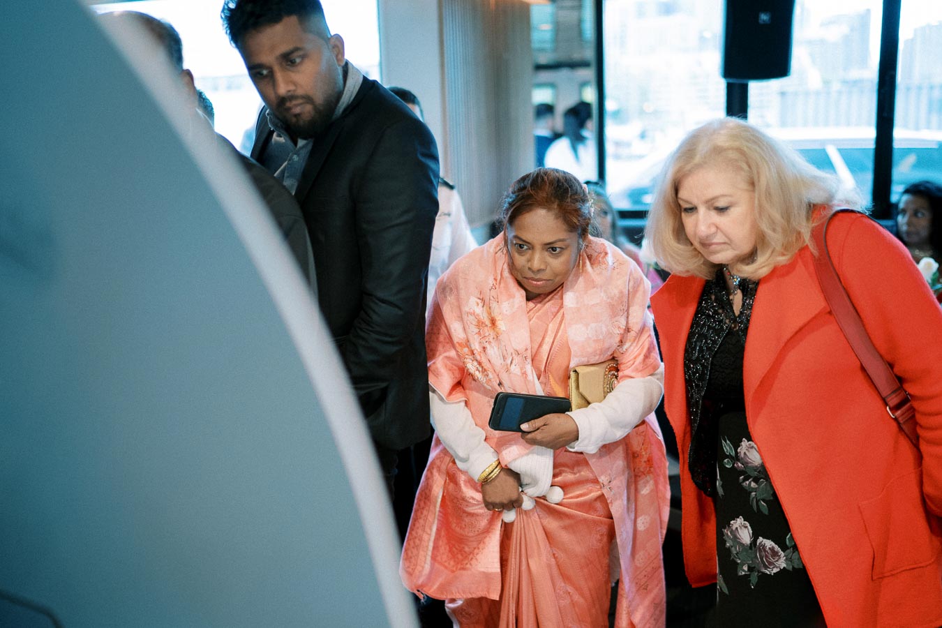 A diverse group of people attentively looking at a display screen at an indoor event, with cityscape visible through the window in the background.