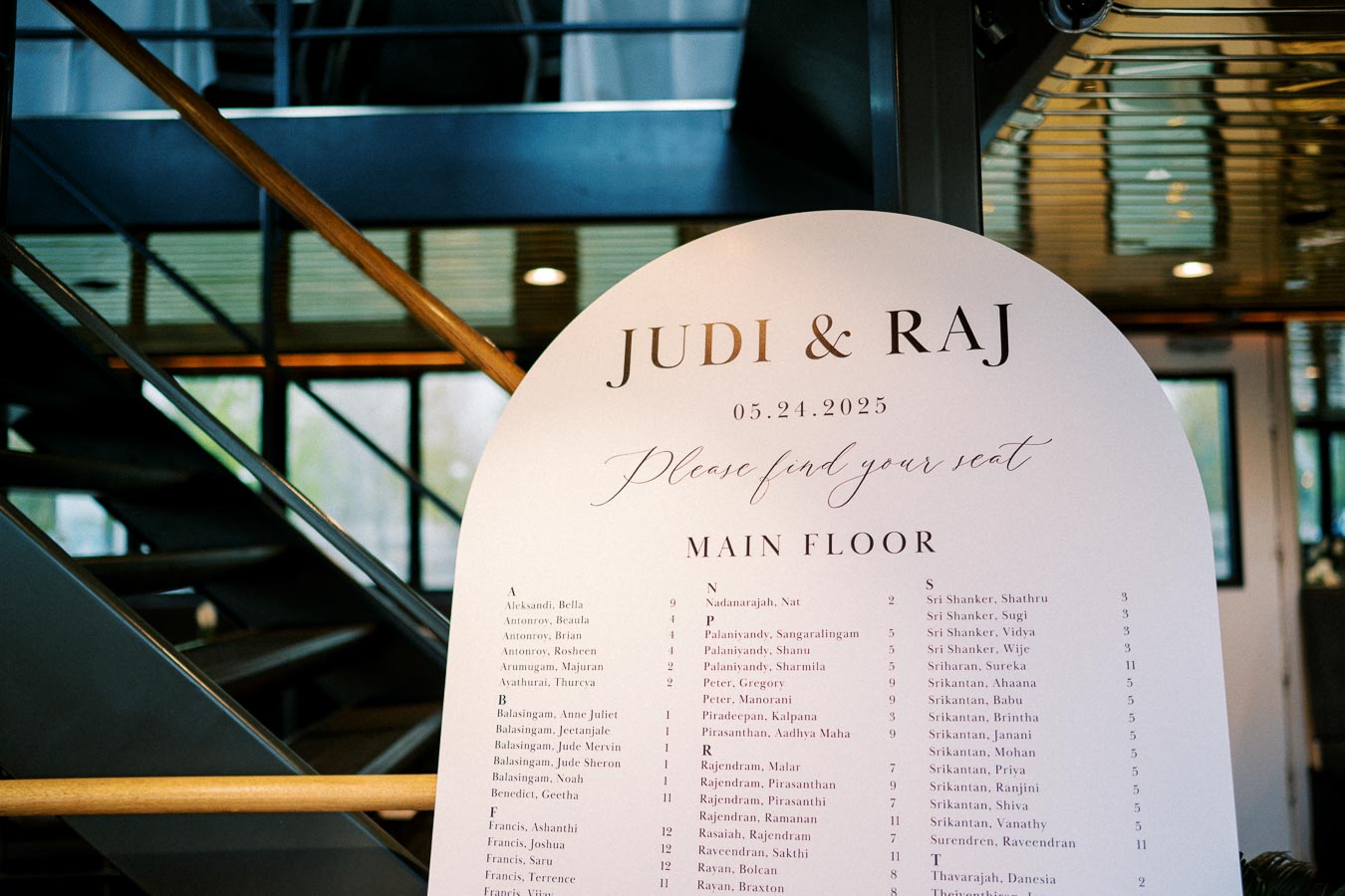 Wedding seating chart for Judi and Raj's event on May 24, 2025, featuring guest names and table assignments, placed in a modern venue with stairs in the background.