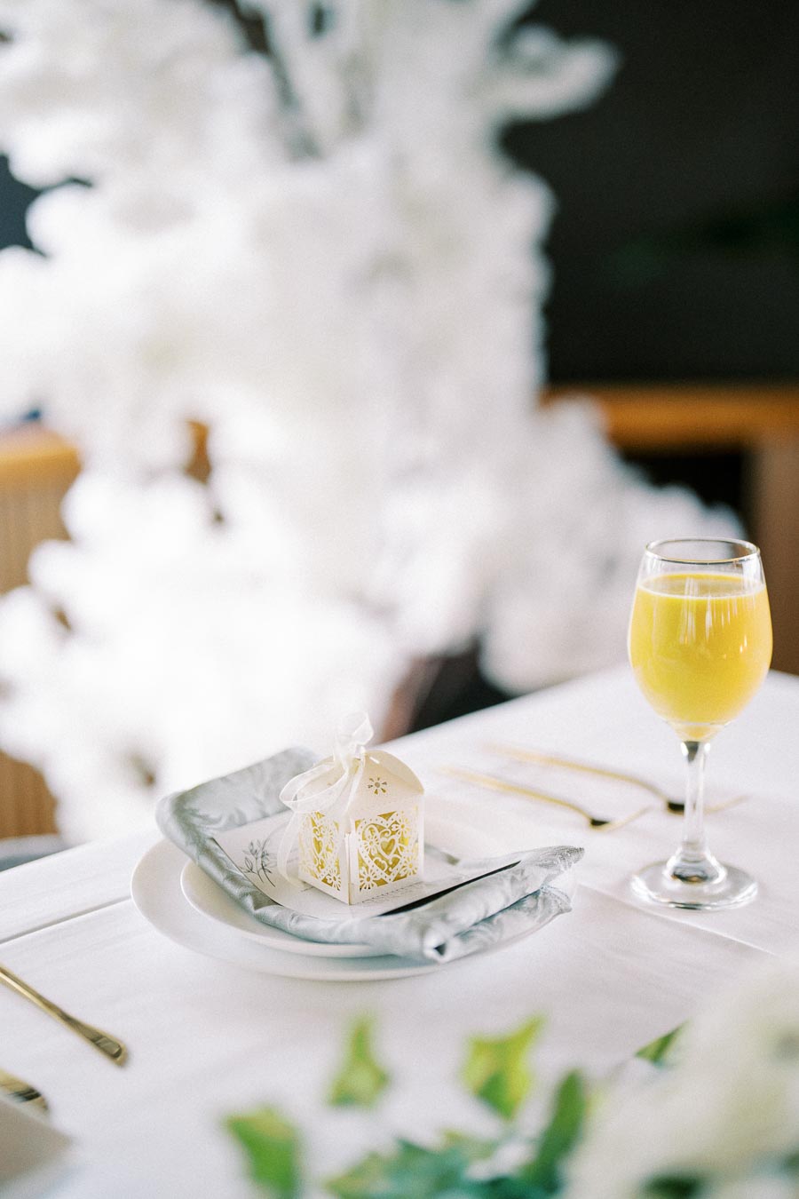 Elegant wedding table setting with a decorative gift box and a glass of orange juice on a white tablecloth, accented by delicate golden cutlery and white floral arrangements in the background.