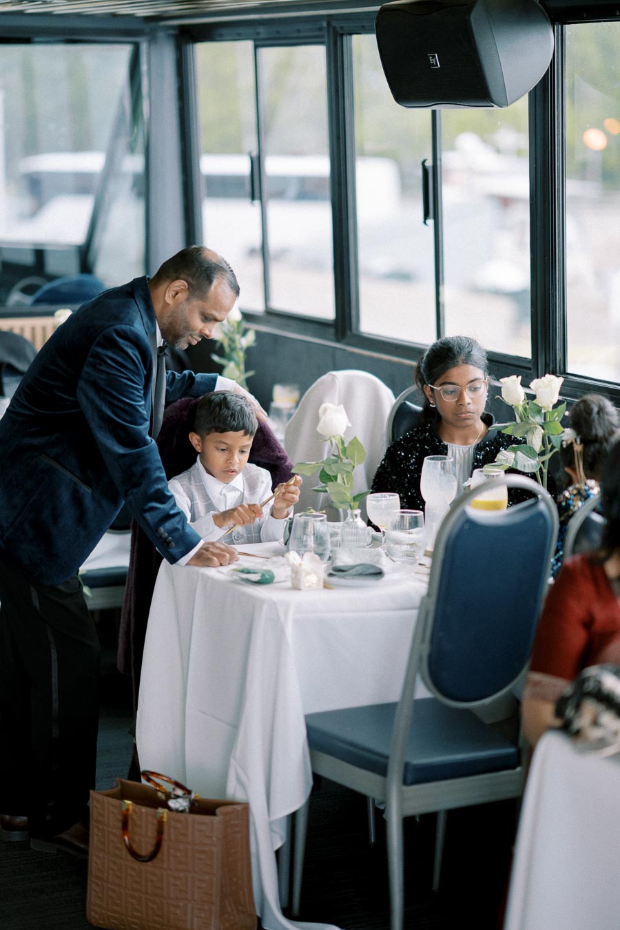 A man in a formal suit assisting a young boy seated at a elegantly set dining table with white tablecloths and fresh flowers. A girl wearing glasses is also at the table, creating a sophisticated and family-friendly dining atmosphere. The setting appears to be on a boat with large windows offering scenic views outside.
