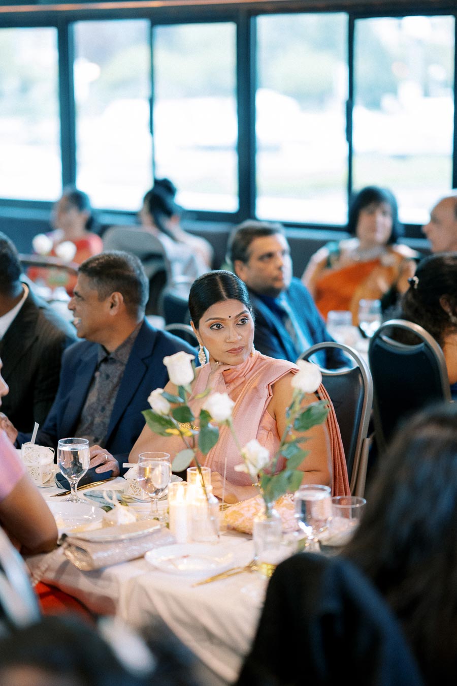 Elegant woman in pink saree attending a formal event, seated at a table with floral centerpiece, surrounded by other guests in a well-lit banquet hall.