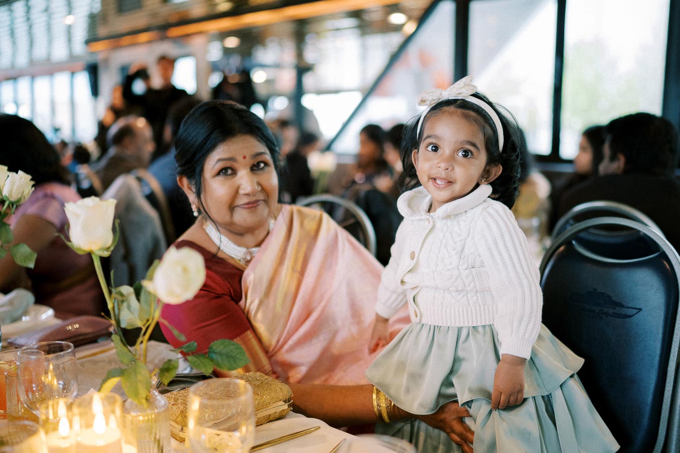 Grandmother and granddaughter enjoying a family gathering at a decorated table, surrounded by guests and warm candlelight, capturing a joyful and affectionate moment.