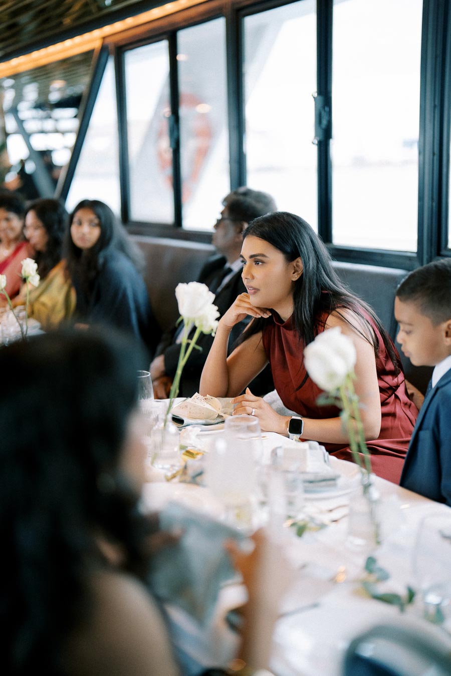 People sitting at a dining table engaged in conversation during an indoor event, with elegant table settings and white roses as centerpiece decorations.
