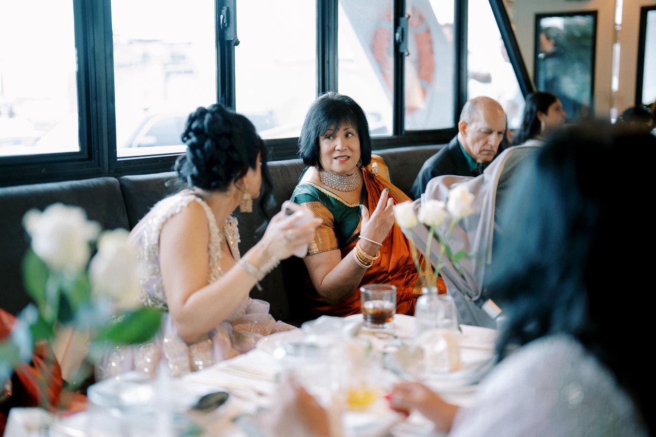 A group of people enjoying a lively discussion at a restaurant table, with vibrant decorations and elegant attire, featuring a woman in an orange and green sari engaging with others.