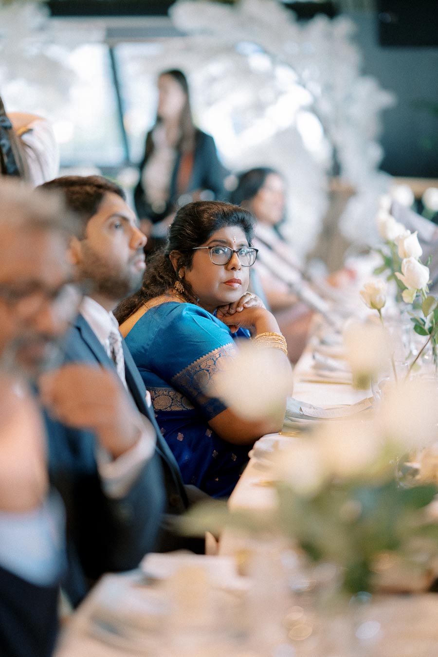 A woman in a blue sari attentively listens during a formal event, surrounded by other attendees seated at a table with white flowers and elegant tableware.