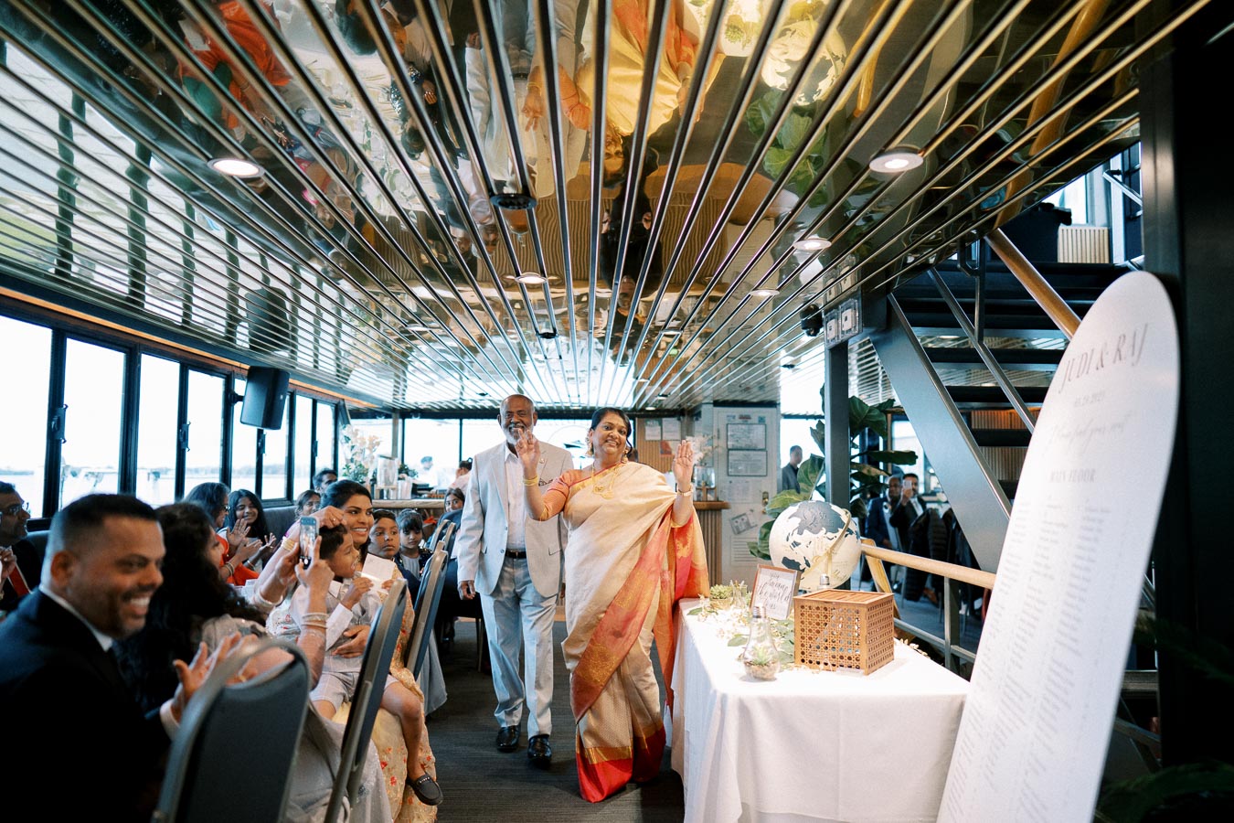 Elegant couple entering a beautifully decorated venue filled with joyful guests, featuring a decorative globe and seating chart, with reflections on the mirrored ceiling, capturing a vibrant celebratory atmosphere.