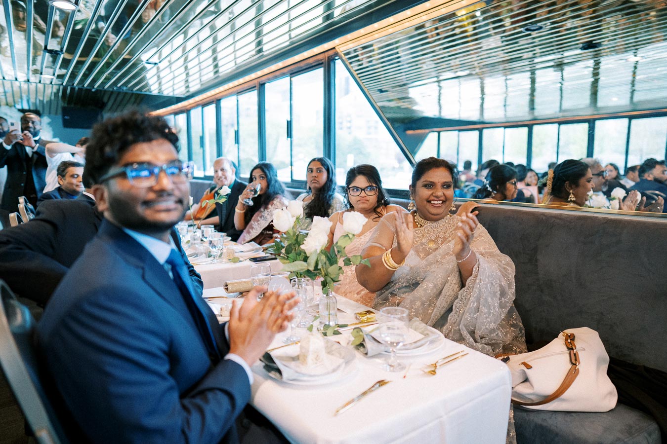 Guests seated at a elegantly decorated table, smiling and clapping during a celebratory event on a boat with large windows and a mirrored ceiling.