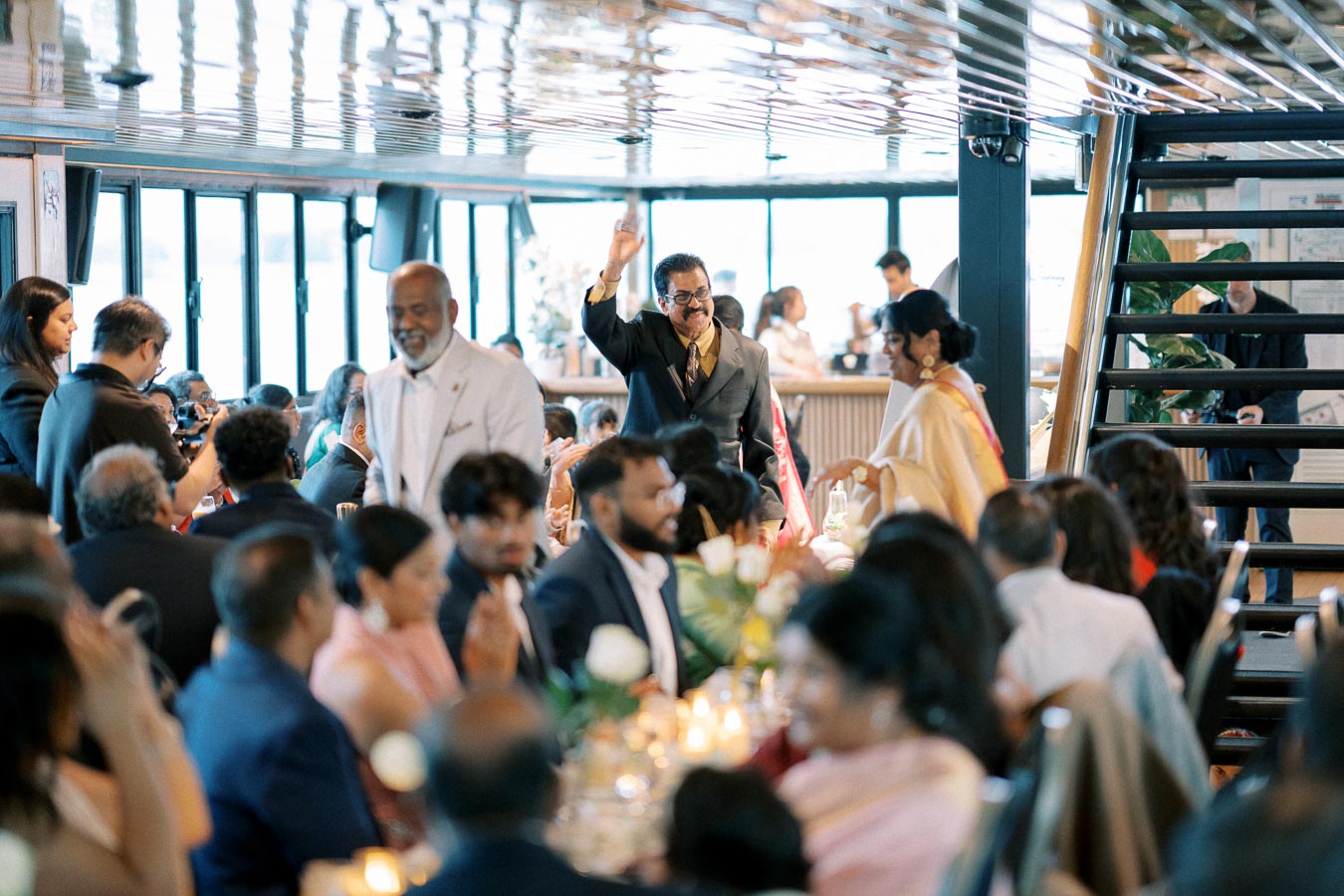 A lively wedding reception on a boat with guests seated at elegantly decorated tables, a man in a suit raising his arm, and staff in the background preparing food.
