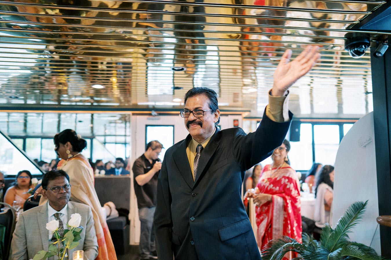 A man in a dark suit waves cheerfully at a festive indoor gathering, with guests seated and mingling around tables adorned with candles and white roses.