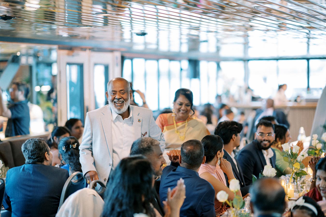 A joyful elderly couple walks through a beautifully decorated dining hall filled with guests during a festive event, surrounded by elegantly arranged tables and floral centerpieces.
