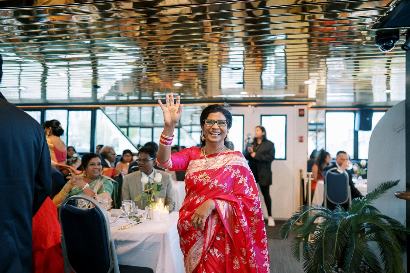 A woman in a vibrant pink and red saree waves cheerfully at a celebration, surrounded by guests seated at elegantly set tables inside a warmly lit venue.
