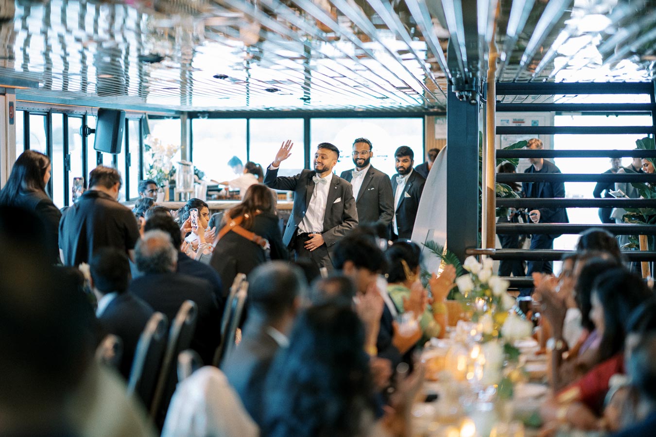 A man in a formal suit waves and enters a bustling indoor event space filled with seated guests, while two others follow him smiling, capturing a warm and celebratory atmosphere.