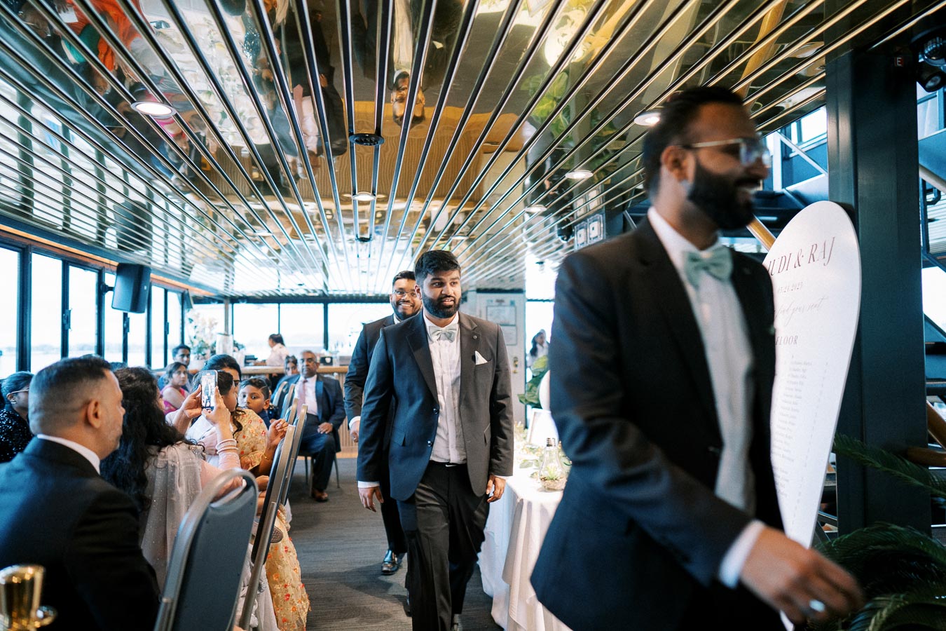 A group of well-dressed people walking down a decorated hallway, possibly at a wedding reception, with guests seated and a decorative mirror ceiling reflecting the event.