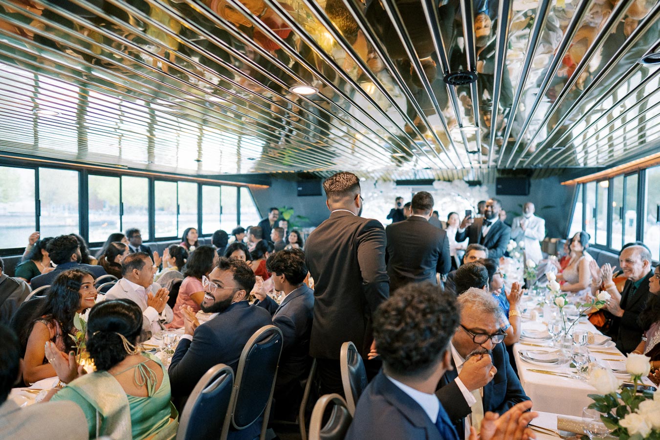 Elegant indoor wedding reception on a luxury boat, featuring well-dressed guests socializing and dining at tables adorned with white tablecloths and floral centerpieces.