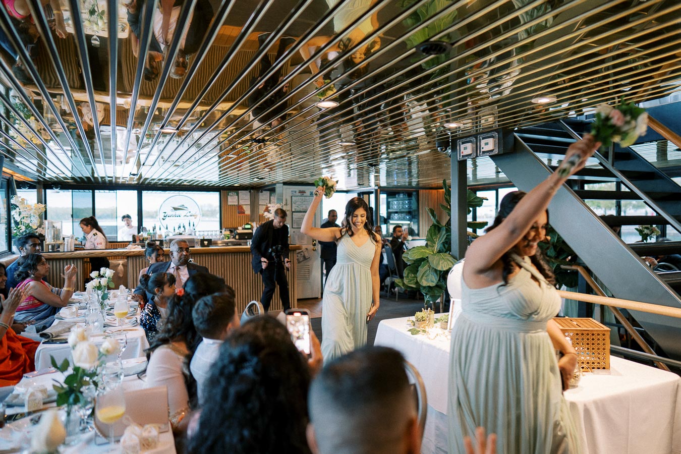 A lively wedding reception on a boat with guests seated at elegantly decorated tables, featuring two bridesmaids wearing light green dresses celebrating with raised arms near a staircase.