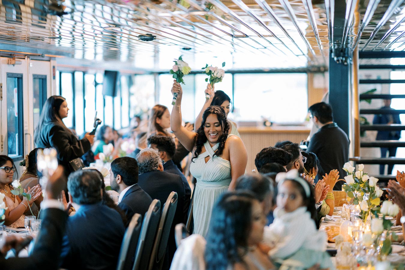 A joyful bridesmaid raises her bouquet while smiling at a lively indoor wedding reception, surrounded by guests seated at elegantly decorated tables.