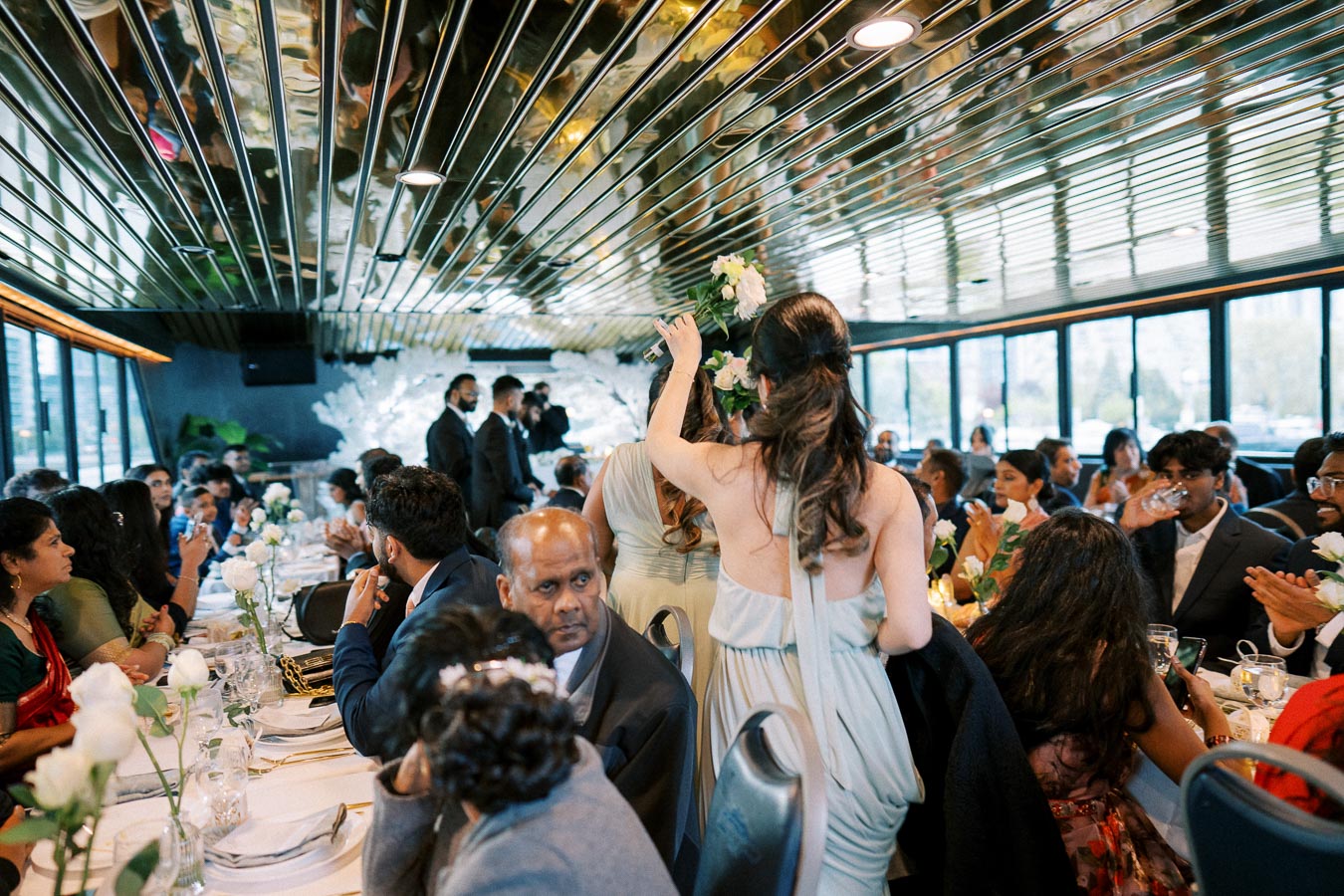 A lively wedding reception with guests seated around elegantly decorated tables, featuring floral centerpieces and ambient lighting. A woman in a light blue dress is walking between the tables holding a bouquet, while guests engage in conversation, creating a festive atmosphere.