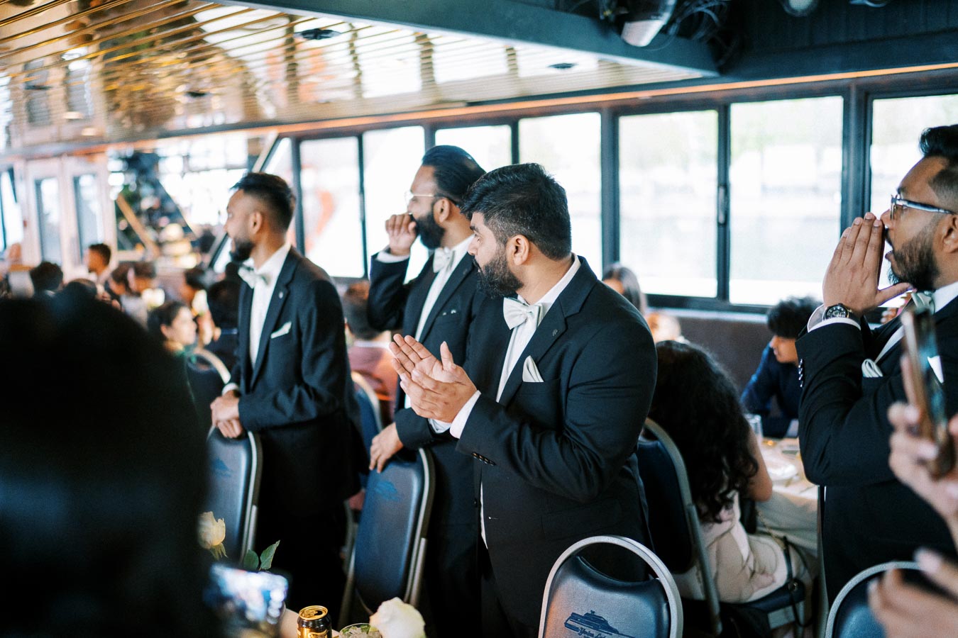 Group of well-dressed men in suits and bow ties clapping and conversing at an elegant indoor gathering or event.
