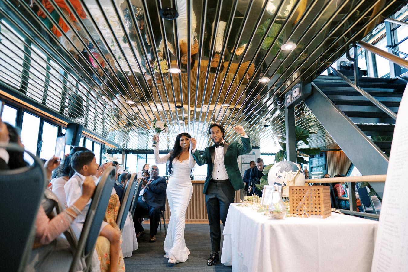 A joyful bride and groom enter a modern wedding reception venue, decorated with elegant wooden and glass elements, while guests applaud and celebrate their entrance.