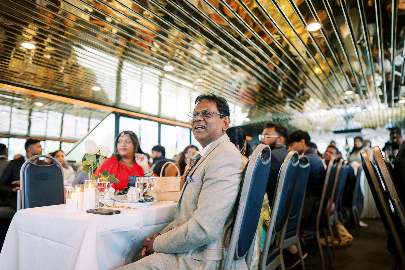Guests at a formal event seated at a table, featuring elegant décor with white roses and candles. The room has a reflective ceiling and large windows, creating a sophisticated ambiance.
