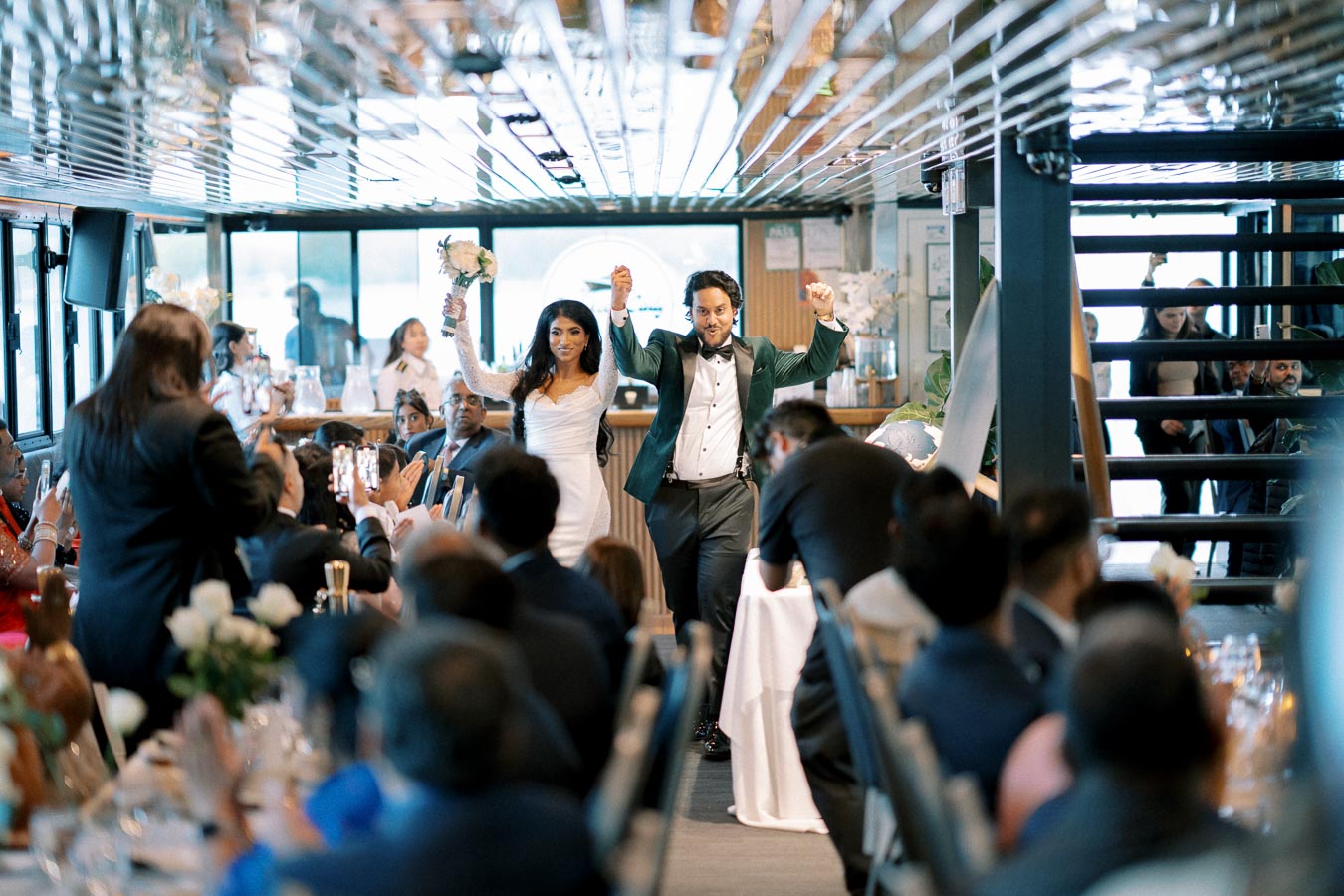A newlywed couple joyfully enters a reception hall, greeted by applauding guests. The bride, holding a bouquet, wears a white dress with lace sleeves, while the groom is dressed in a stylish tuxedo. The venue is elegantly decorated, and guests are capturing the moment on their phones.