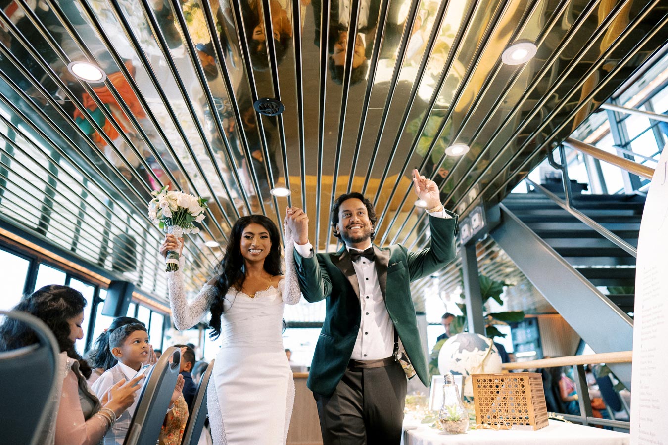 Bride and groom joyfully enter a wedding venue, with the bride in a white dress holding a bouquet and the groom in a green suit jacket, surrounded by smiling guests, modern decor, and reflective ceiling panels.