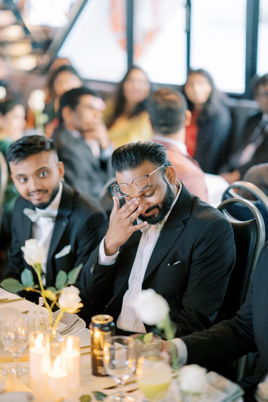 A man wearing glasses and a black suit sits at a formal event table, smiling and touching his face, while another man in a bow tie looks on. The table is elegantly set with candles and white roses, surrounded by other seated guests in a softly blurred background.