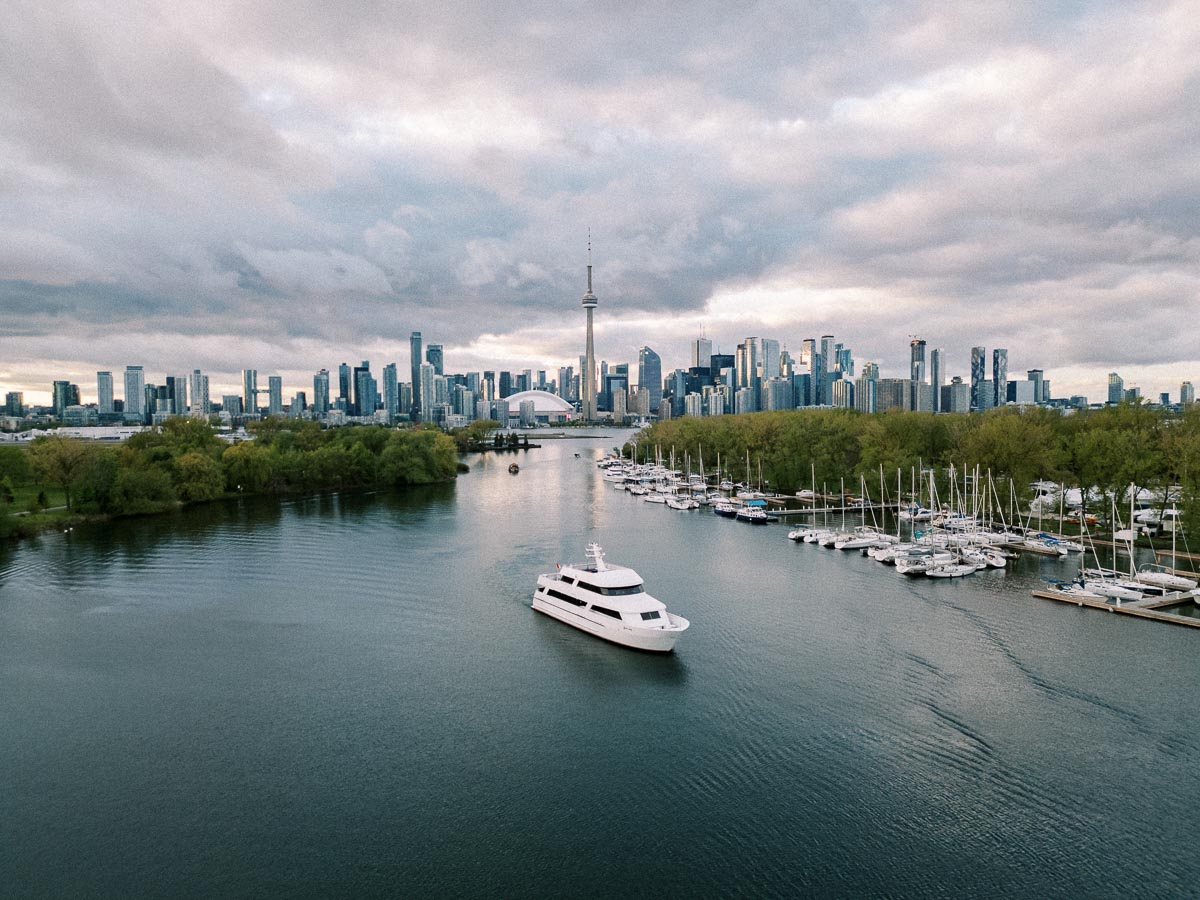 A white yacht cruising through a body of water with a marina full of boats to the right and the Toronto skyline, featuring the CN Tower, under a cloudy sky in the background.
