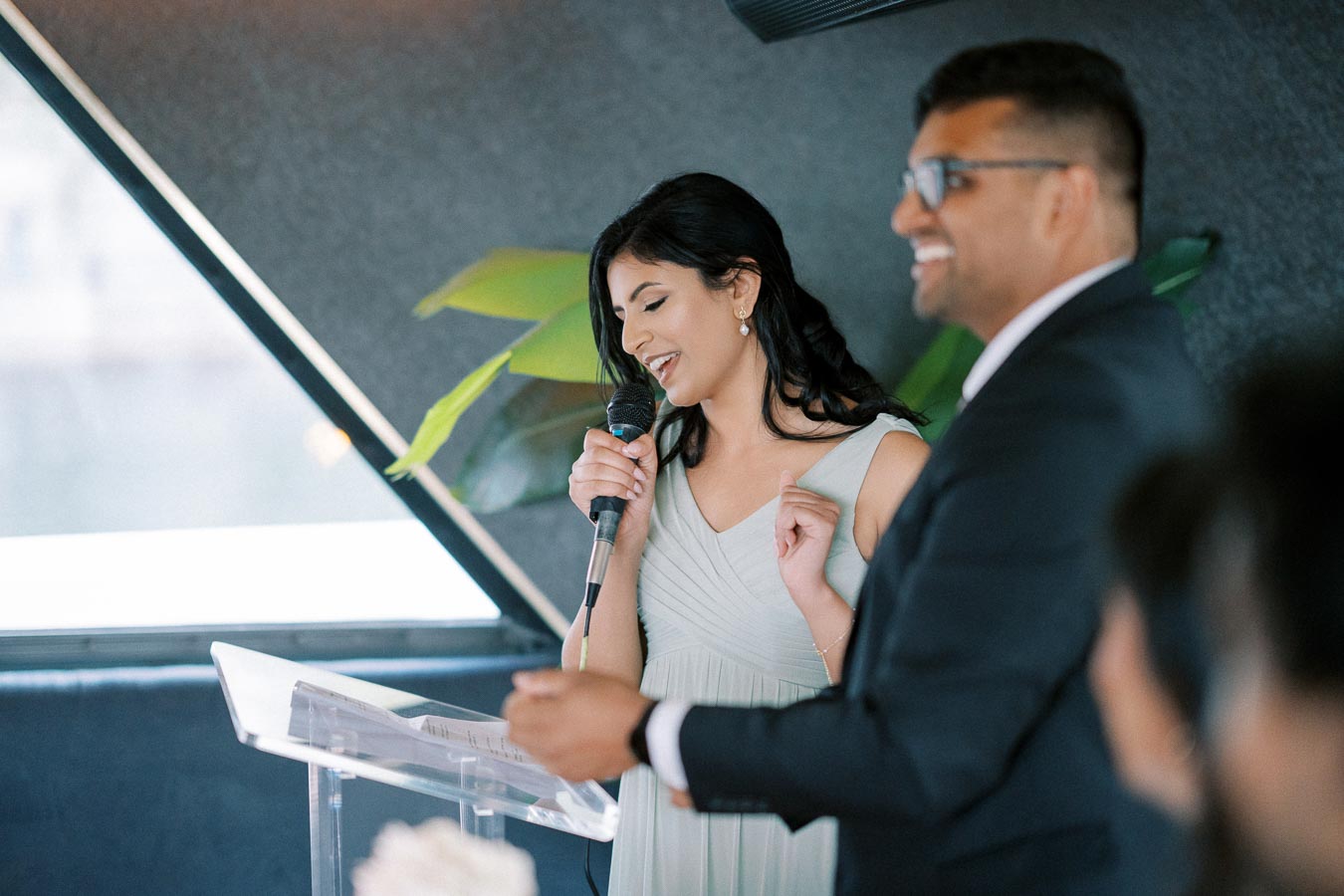 Two people smiling and speaking at an event podium, with the woman holding a microphone and enjoying the moment, while a blurred background shows greenery and interior lighting.