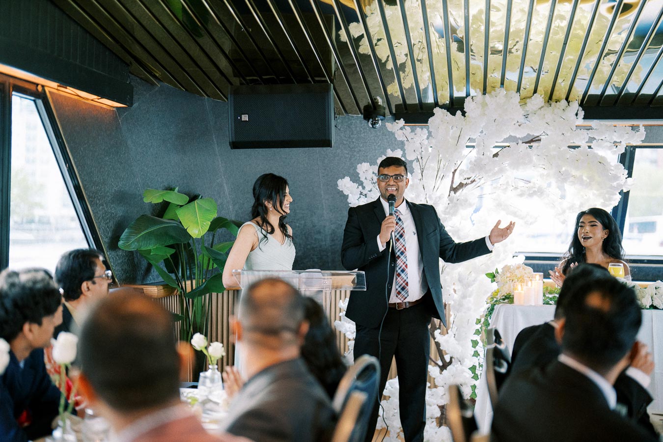 A man in a suit speaks into a microphone during a formal event, with a woman standing beside him and another seated at a decorated table. The room features elegant decor with white flowers and green plants, suggesting a celebration or wedding reception. People in formal attire are seated in the foreground.