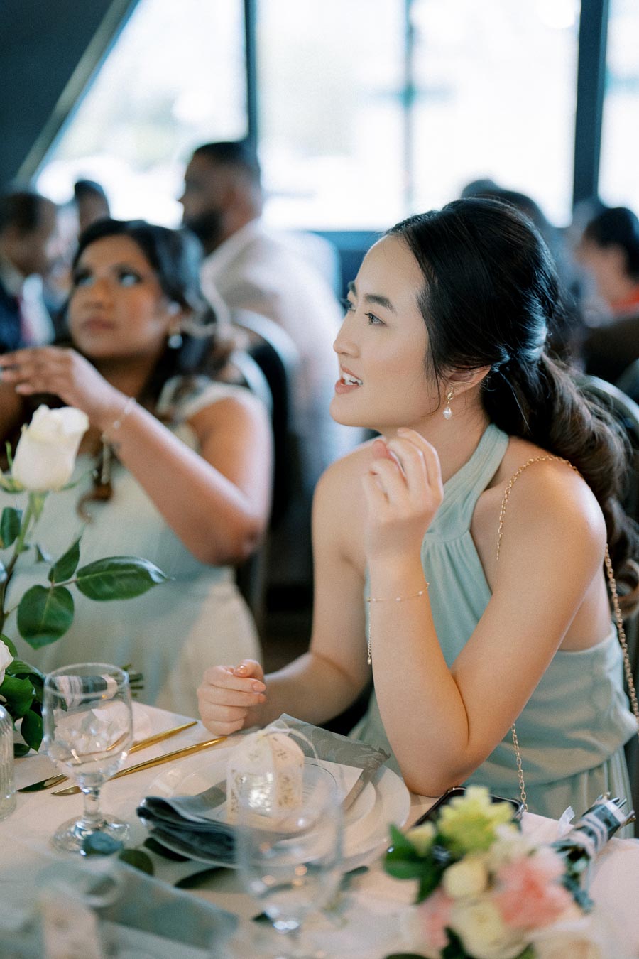 A woman in a light blue dress sitting at a decorated table during a formal event, with a bouquet of flowers and dinnerware, engaging in conversation with surrounding guests.