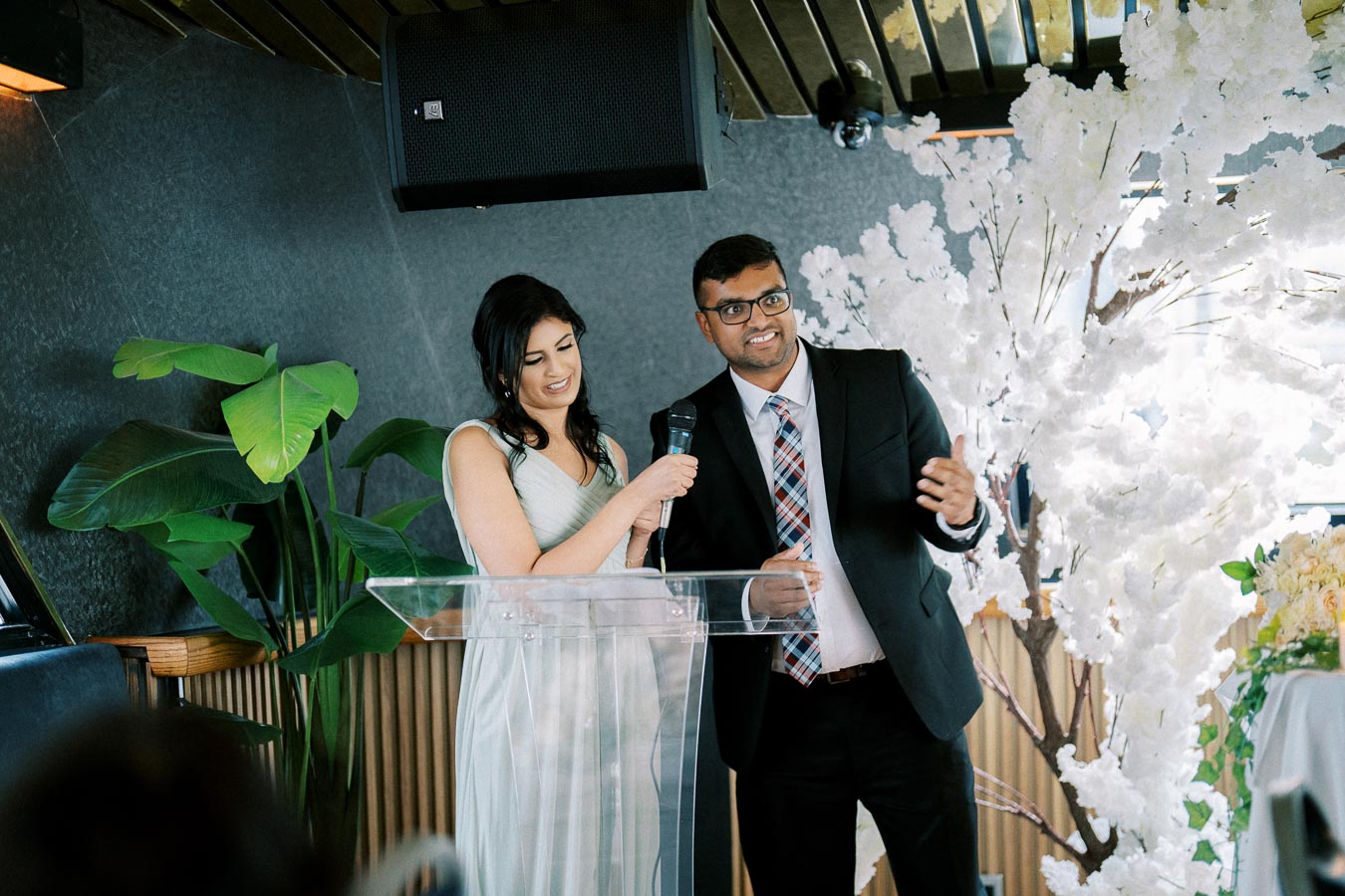 A couple standing at a clear podium, smiling and sharing a microphone, with a backdrop of white floral decorations and greenery, dressed in formal attire.