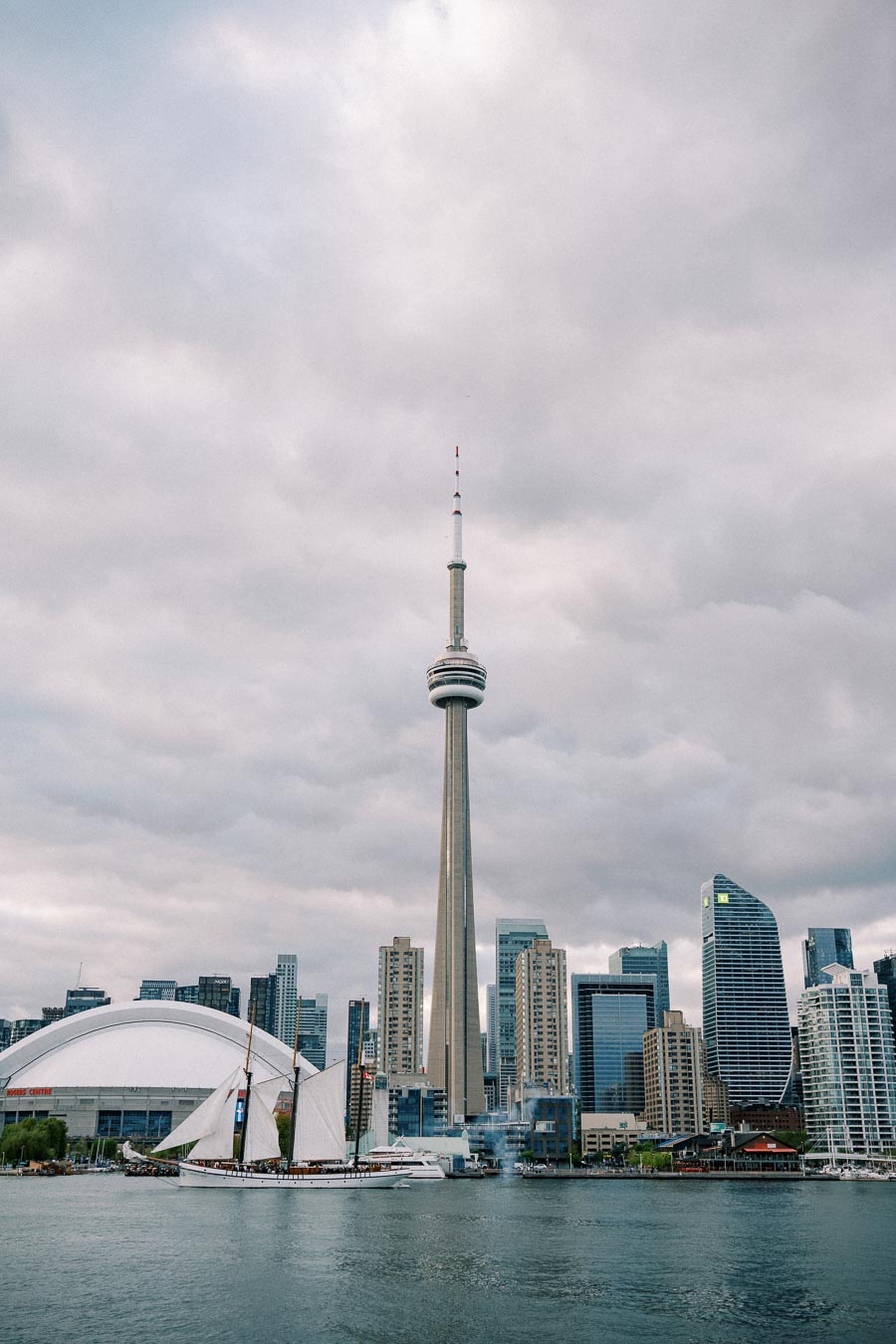 Toronto skyline featuring CN Tower, Rogers Centre, and sailboat on Lake Ontario under cloudy sky