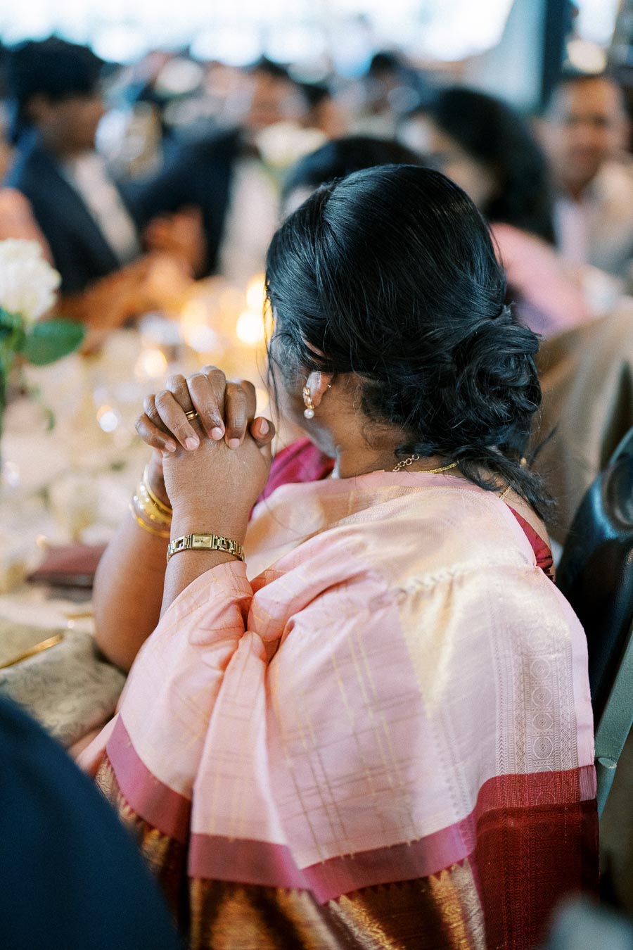 A woman in a colorful traditional sari with intricate designs sits at a dining table during an event, her hands clasped together. She wears a gold watch, earrings, and bracelets. The background features elegantly blurred guests and softly glowing candles, creating a warm, celebratory atmosphere.