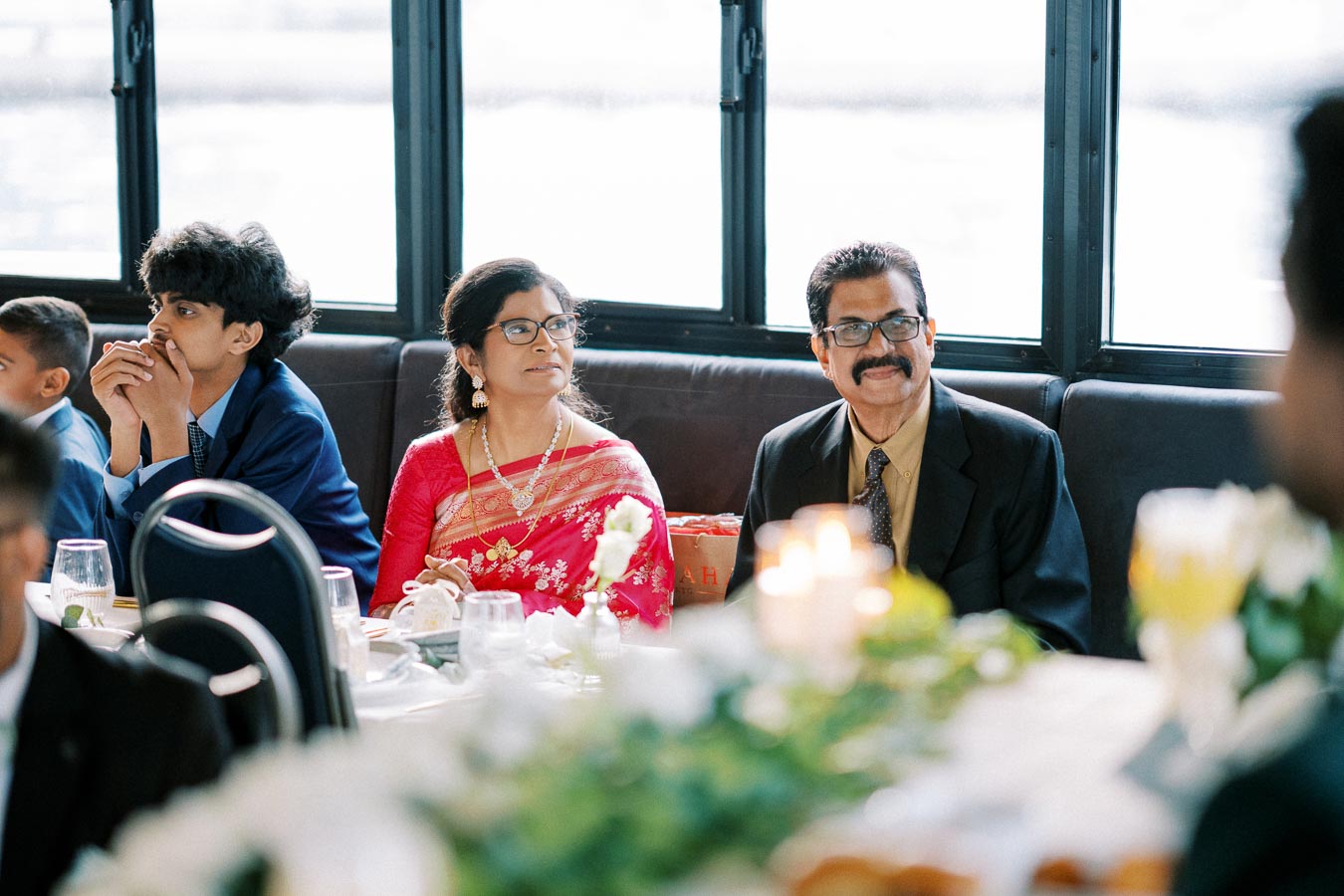 A group of people sit at a beautifully decorated table during a formal event, with a woman in a red saree and a man in a suit, both wearing glasses, attentively listening. The setting is indoors with a window in the background, displaying a bright atmosphere.