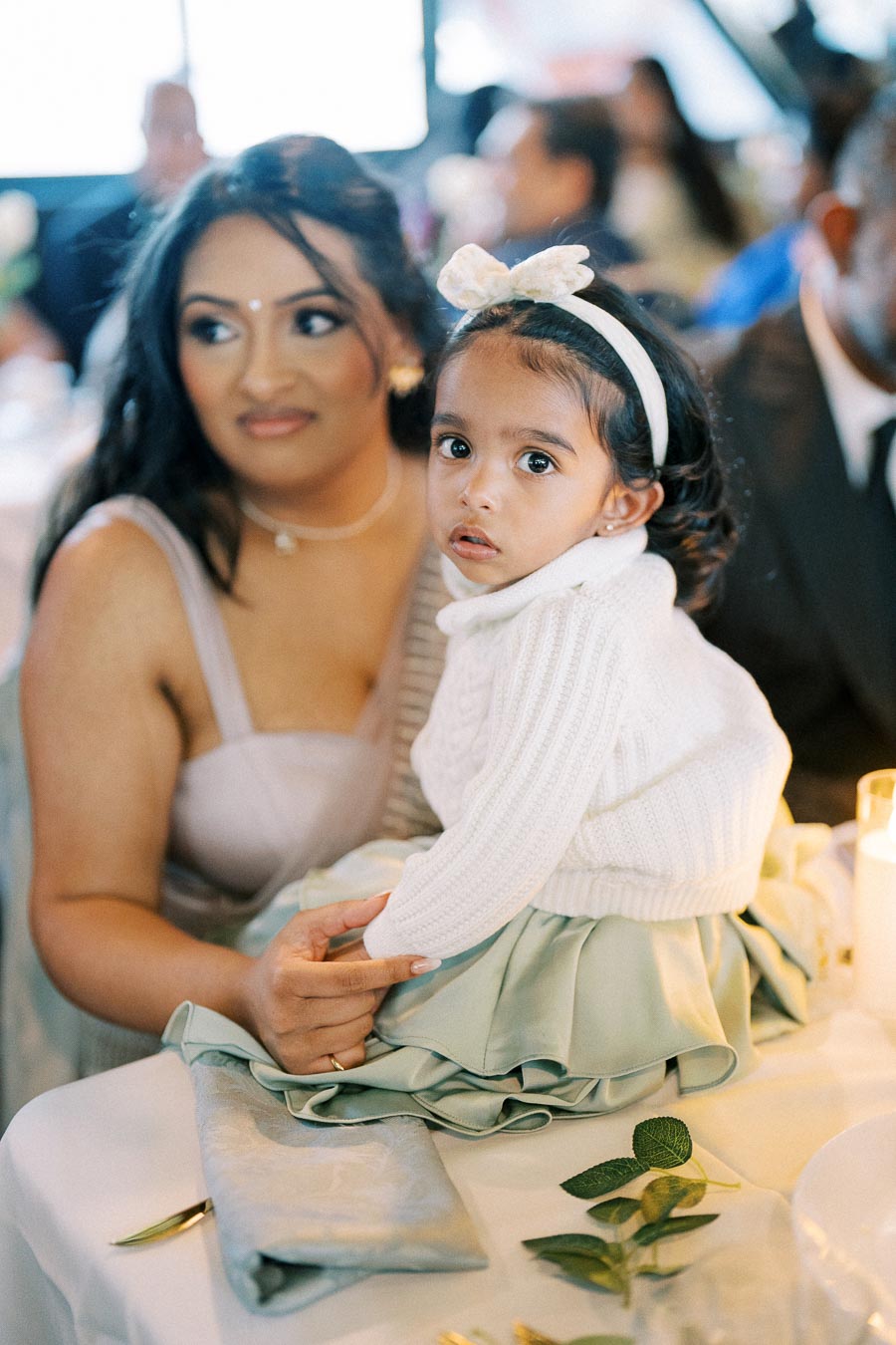 A young girl in a white sweater and headband sits on a woman's lap at a festive event, surrounded by soft lighting and elegant decor.