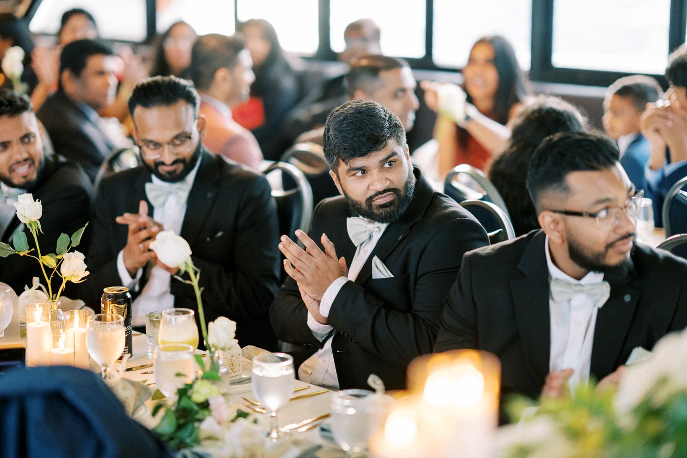 Group of well-dressed men clapping at a formal event, seated at a decorated table with flowers and candles, surrounded by other guests in a celebratory atmosphere.