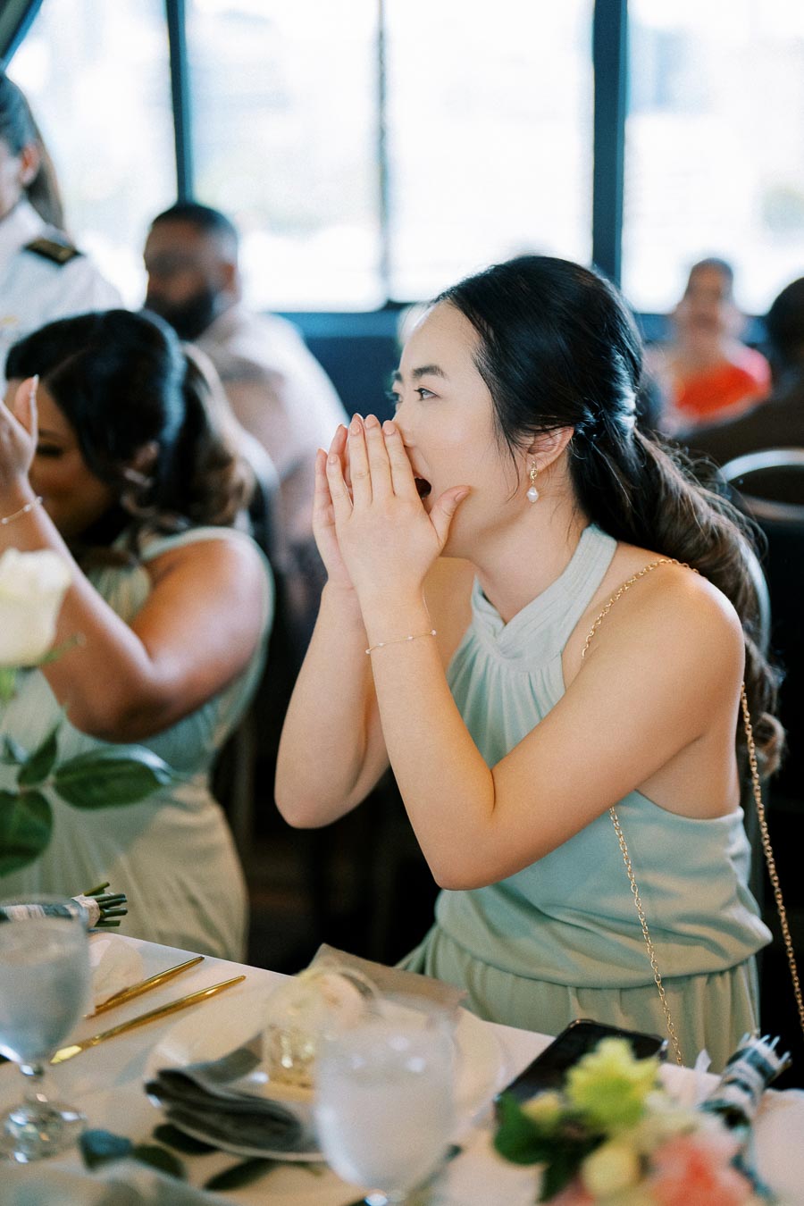 A woman in a light blue dress expressing excitement or surprise at a formal event, seated at a decorated table with flowers and formal dining settings.