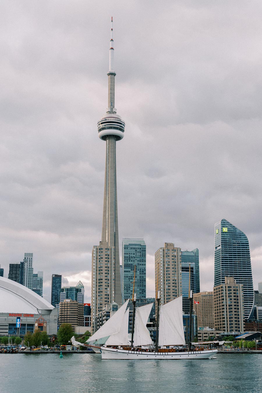 Scenic view of Toronto skyline featuring the CN Tower and a sailboat on Lake Ontario with a cloudy sky backdrop.