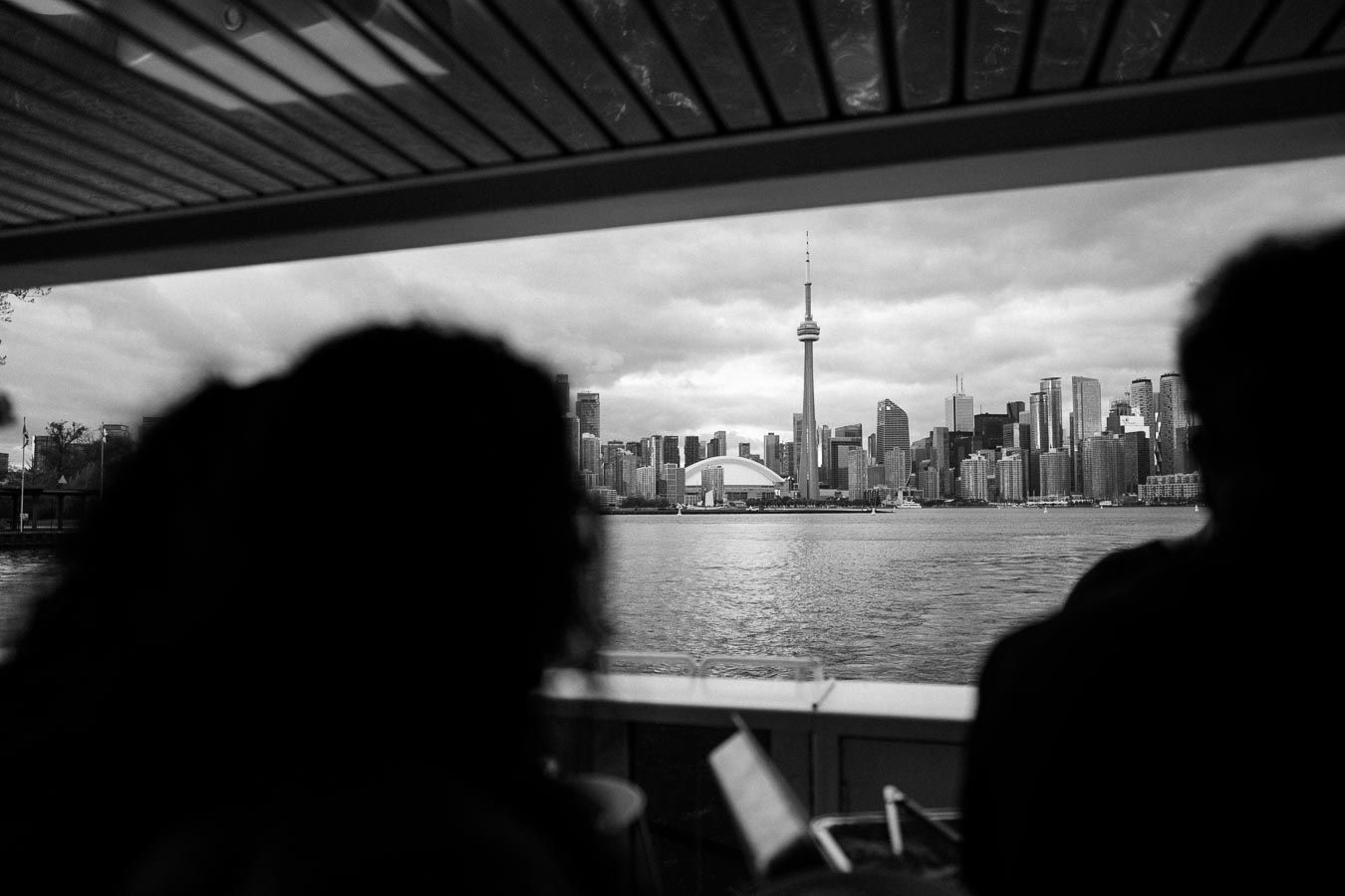 Black and white photo of Toronto's skyline viewed from a boat, featuring the CN Tower and Rogers Centre across Lake Ontario, with silhouettes of people in the foreground.